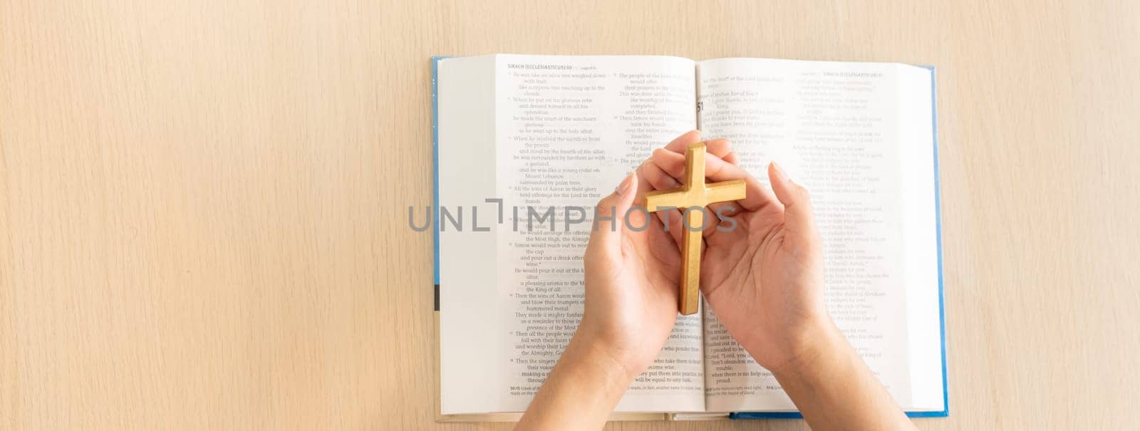 Female god believer holding wooden cross on opened holy bible book at light wooden church table. Top view. Concept of hope, religion, faith, christianity and god blessing. Warm background. Burgeoning.