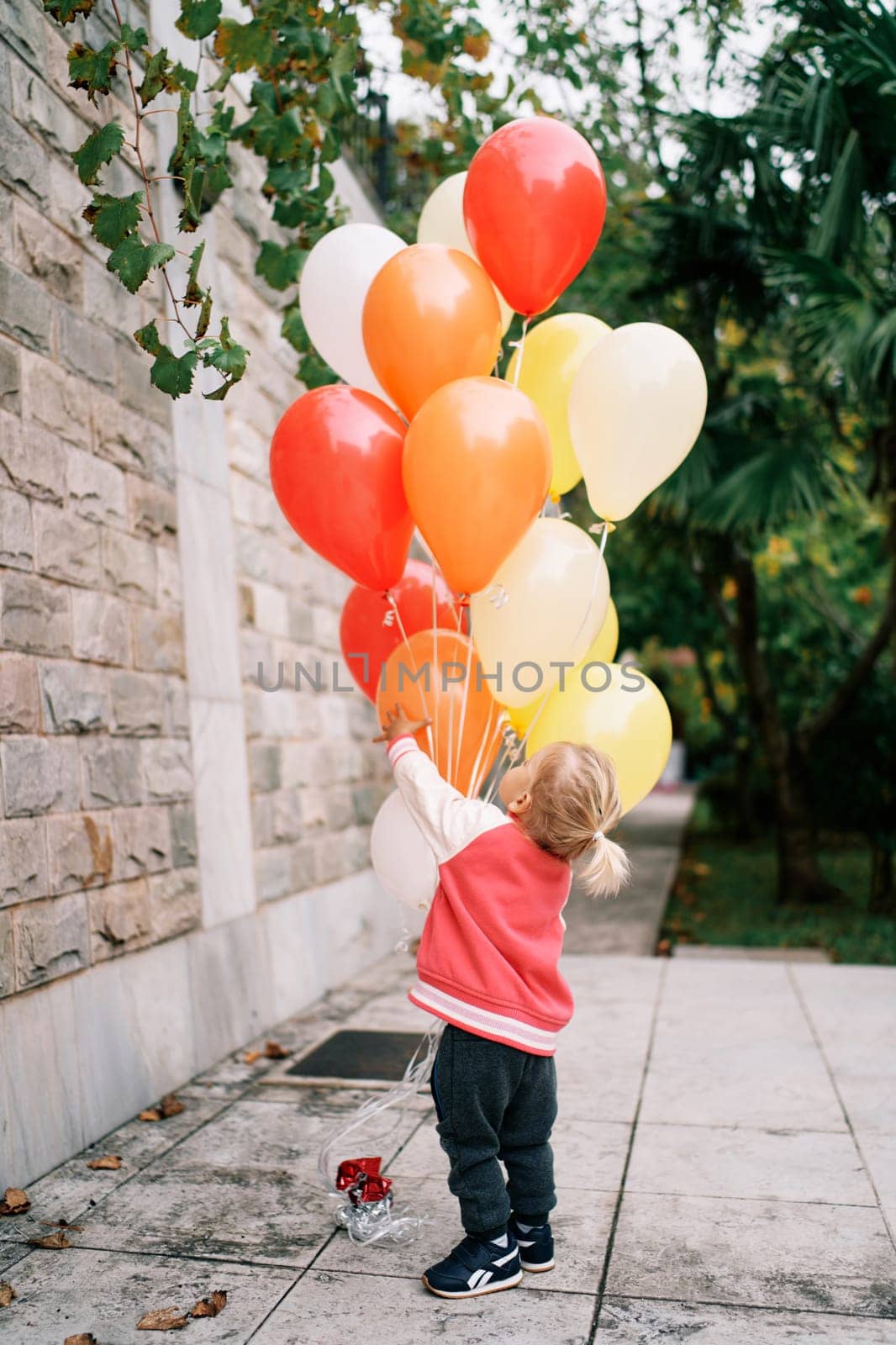 Little girl hugs a bunch of balloons in the garden and looks up. Back view. High quality photo