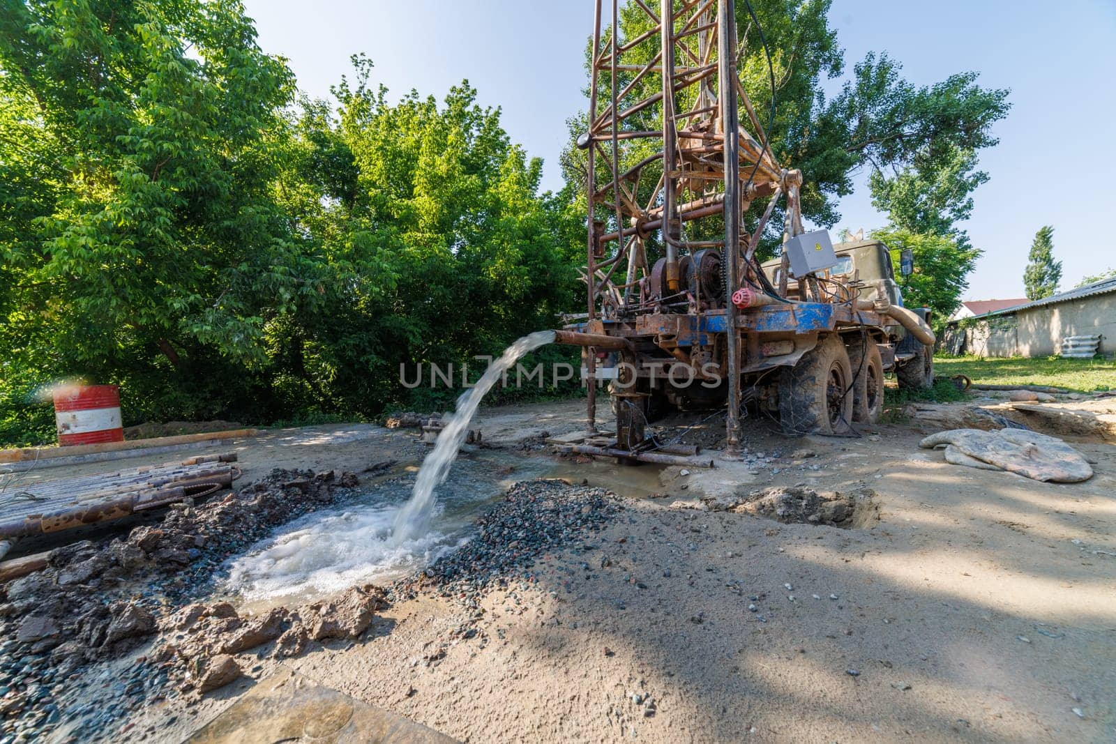 Old water pump truck extracts water from a hole in the ground at sunny summer day, surrounded by a natural landscape with trees, plants, and soil. Ultra-wide angle.