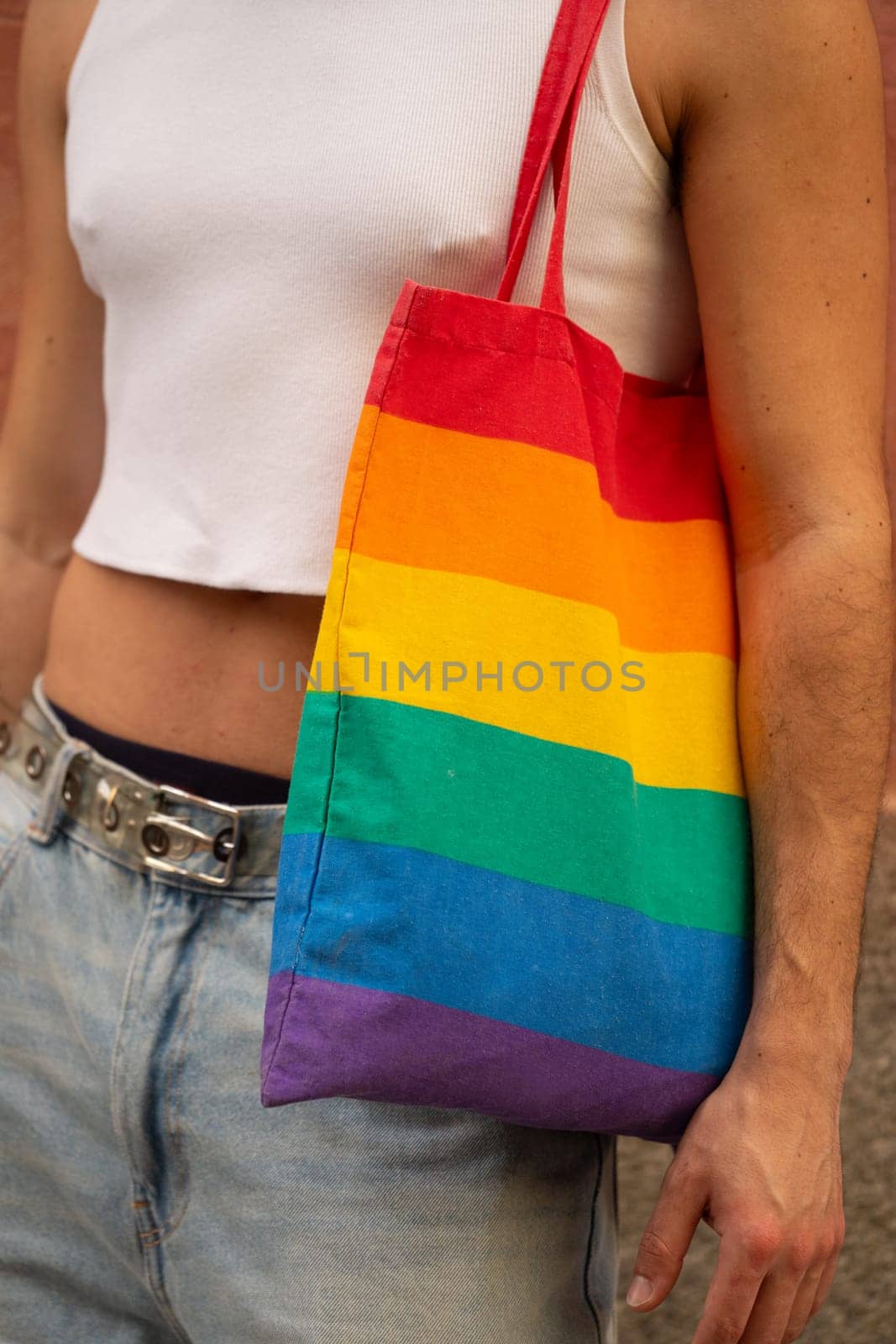 A man is holding a rainbow tote bag.The man is wearing a white shirt and blue jeans. Concept of pride and inclusivity by papatonic