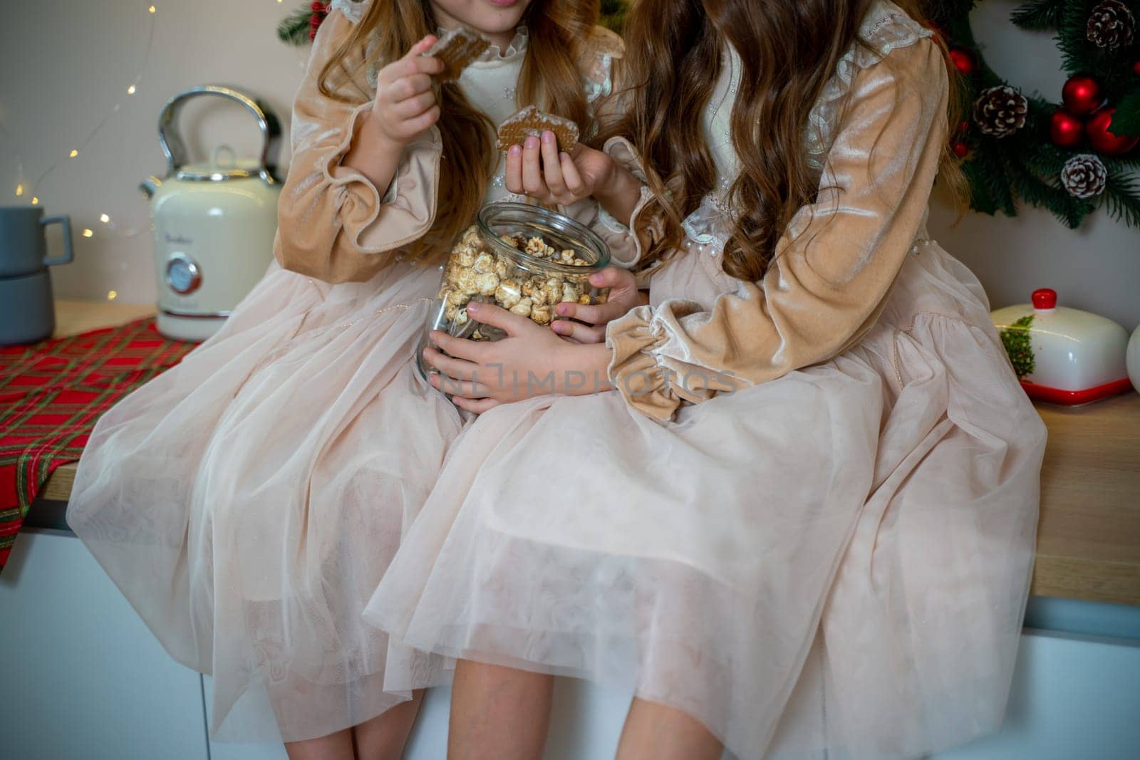 Two cute little girls in beige dresses are sitting on the table holding cookies. New Year decoration. Luxurious New Year's interior