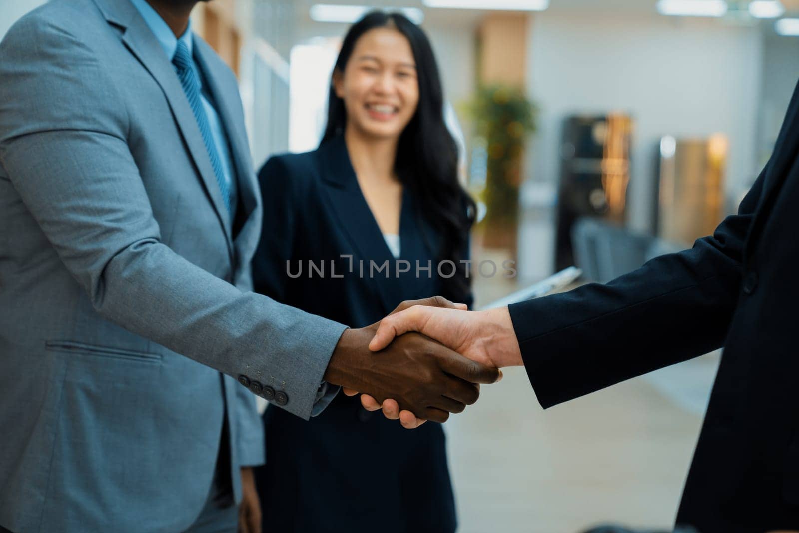 Closeup of business hands shaking between businessman and professional male leader while smart businesswoman stand near at modern office corridor. Represented unity, corporate, teamwork. Ornamented.