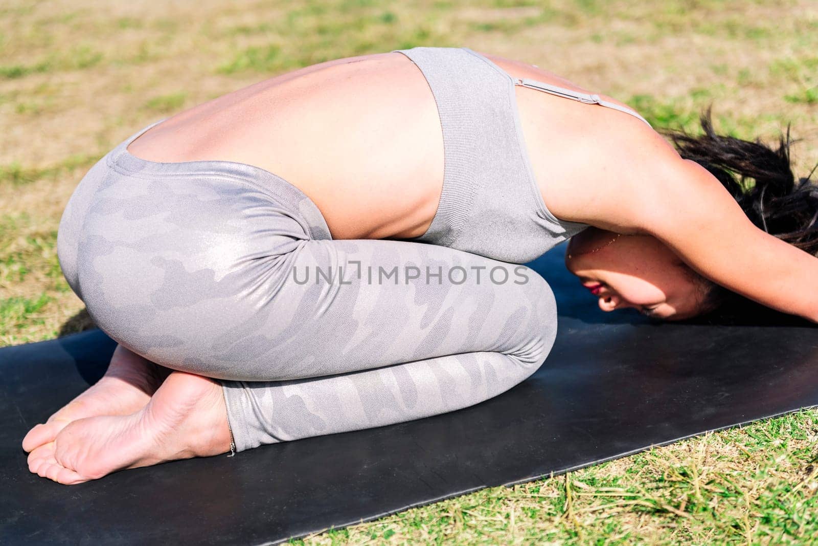 young woman in sportswear kneeling on her yoga mat doing back stretching exercises on the grass in the park, active and healthy lifestyle concept