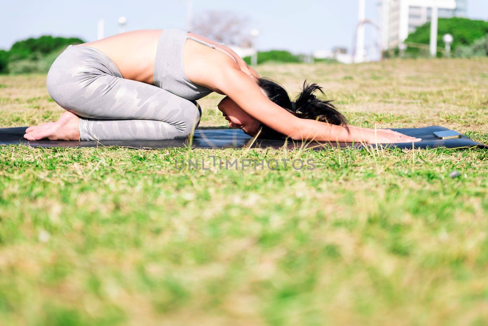 young woman in sportswear kneeling on her yoga mat doing back stretching exercises in the park following the instructions of the app on her mobile phone, active and healthy lifestyle concept