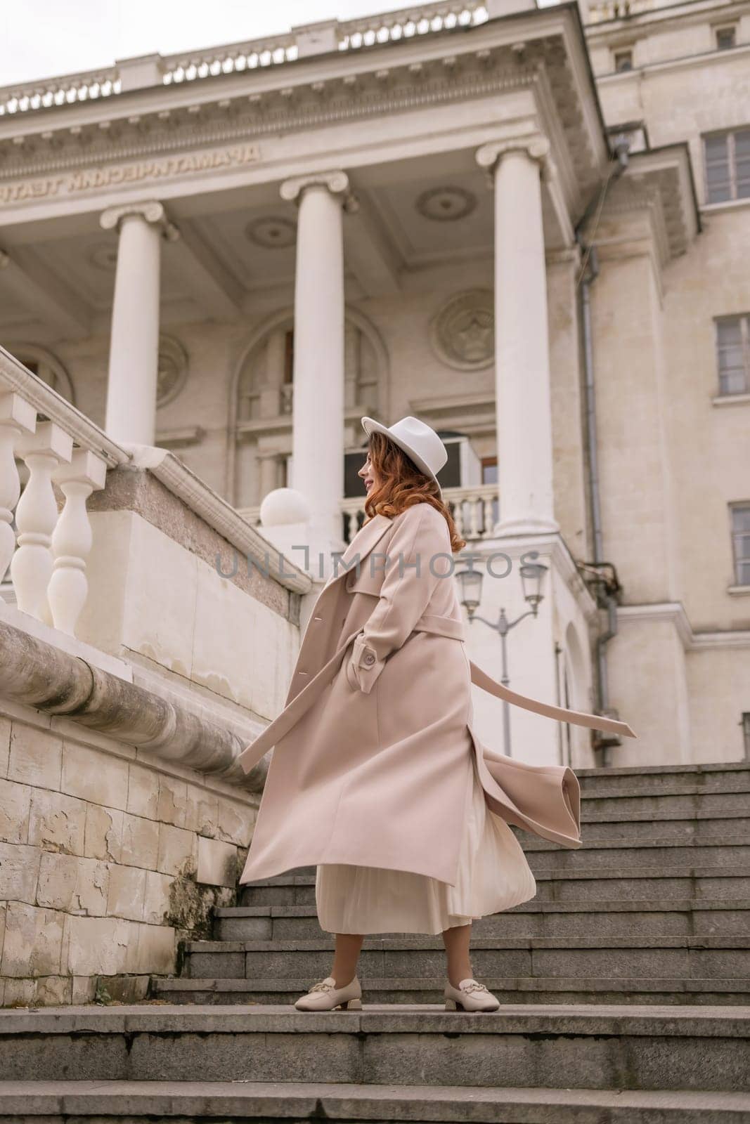 woman in elegant coat and hat against an intricate architectural backdrop, harmoniously blending modern fashion with historical allure. The soft daylight adds to its timeless appeal. by Matiunina