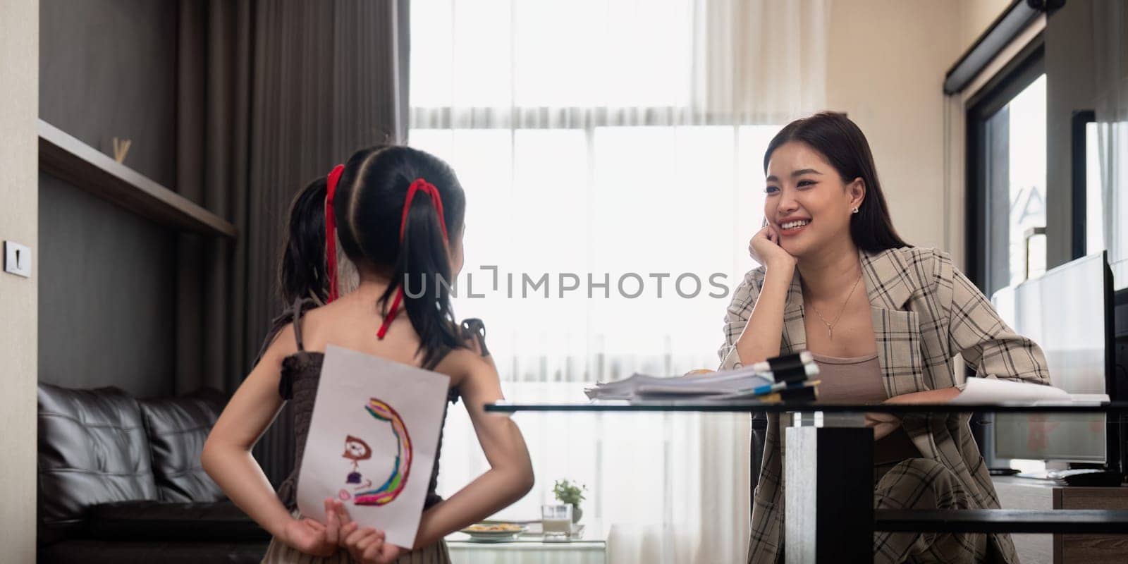 Happy family, mother and little daughter hold drawings for mom to see, sitting in cozy room at home. Working mom work from home office.