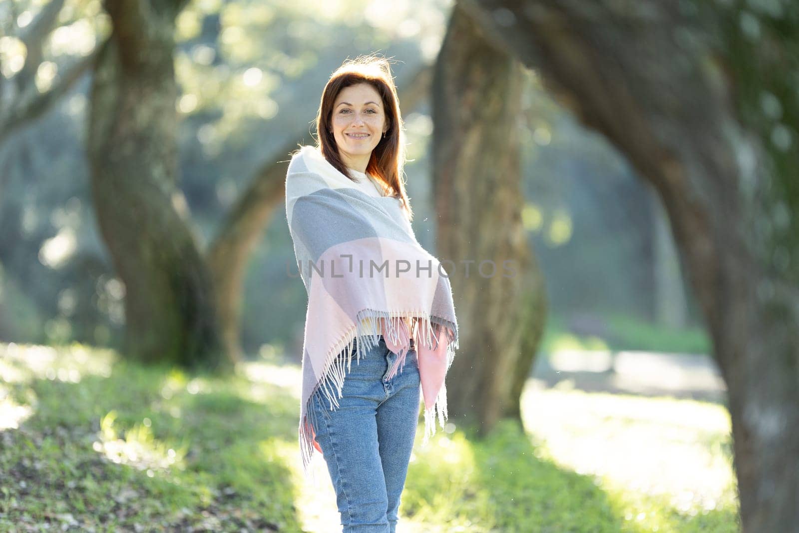 A woman is standing in a park, wearing a colorful scarf and jeans. She is smiling and looking towards the camera