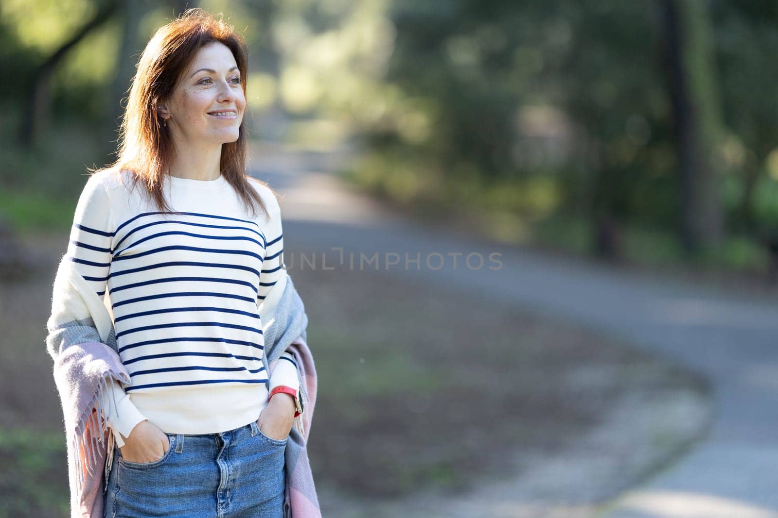 An adult woman with dentistry braces wearing a striped sweater and jeans is standing on a path. She is smiling and looking to her right