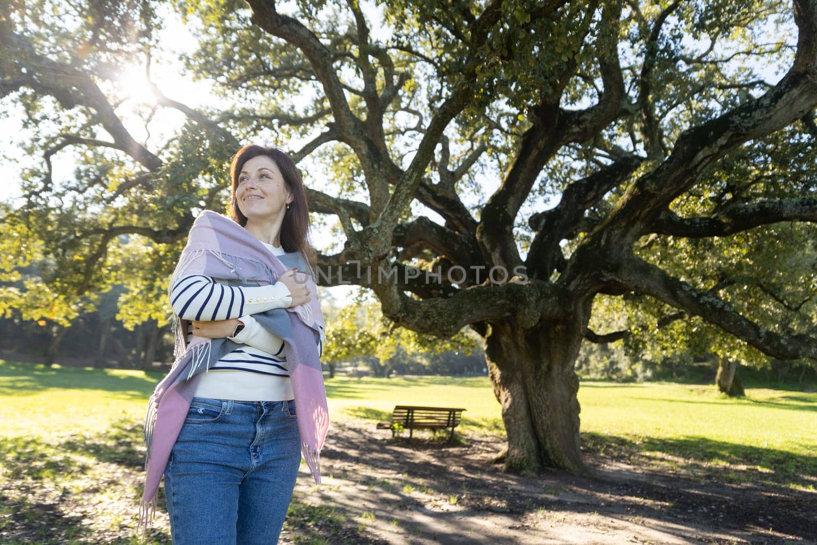 A woman is standing in a park, wearing a scarf and a white shirt. She is smiling and looking up at the sky. The park is filled with trees, and there is a bench nearby