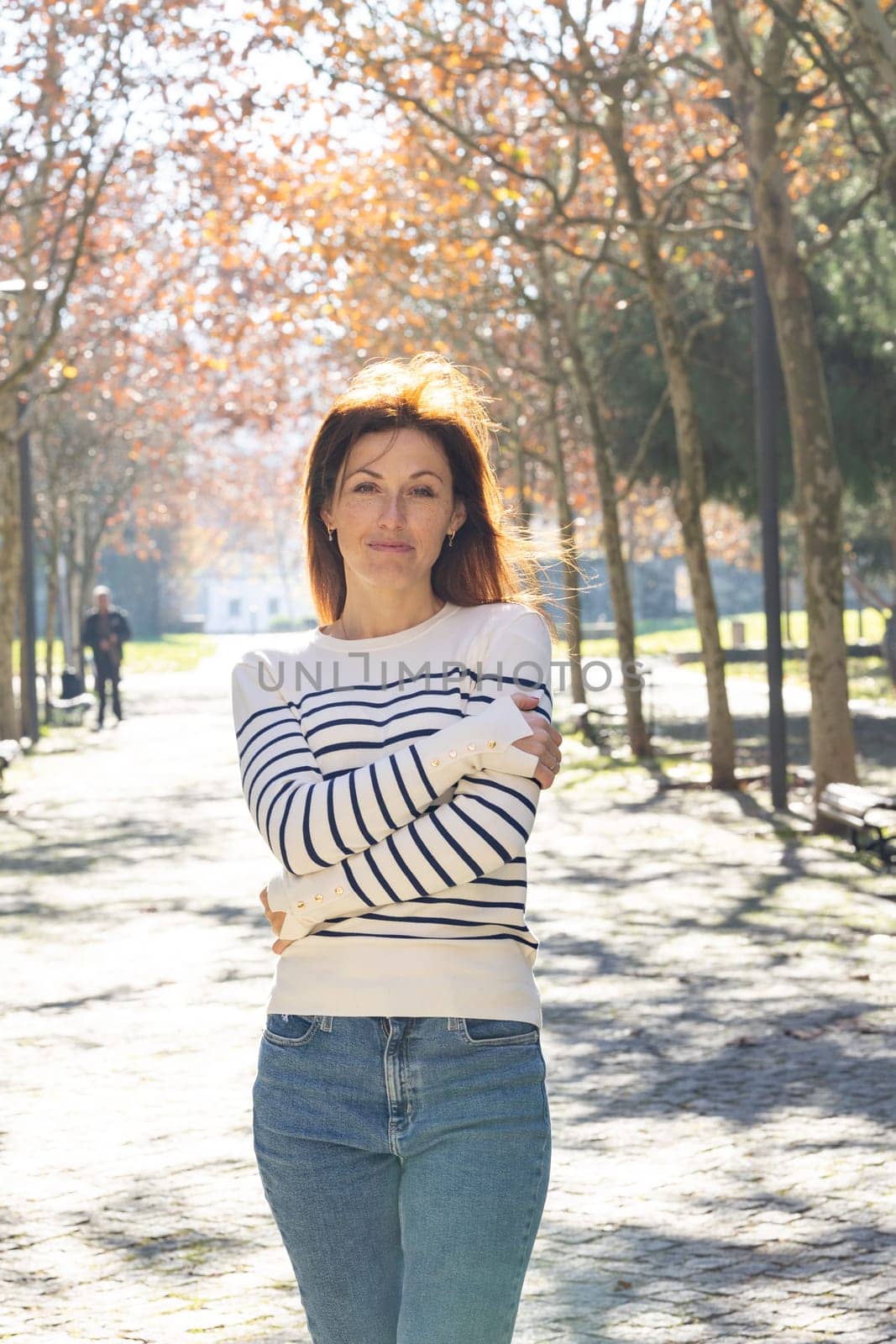 A woman wearing a striped sweater and jeans stands in a park. She is smiling and she is enjoying the day