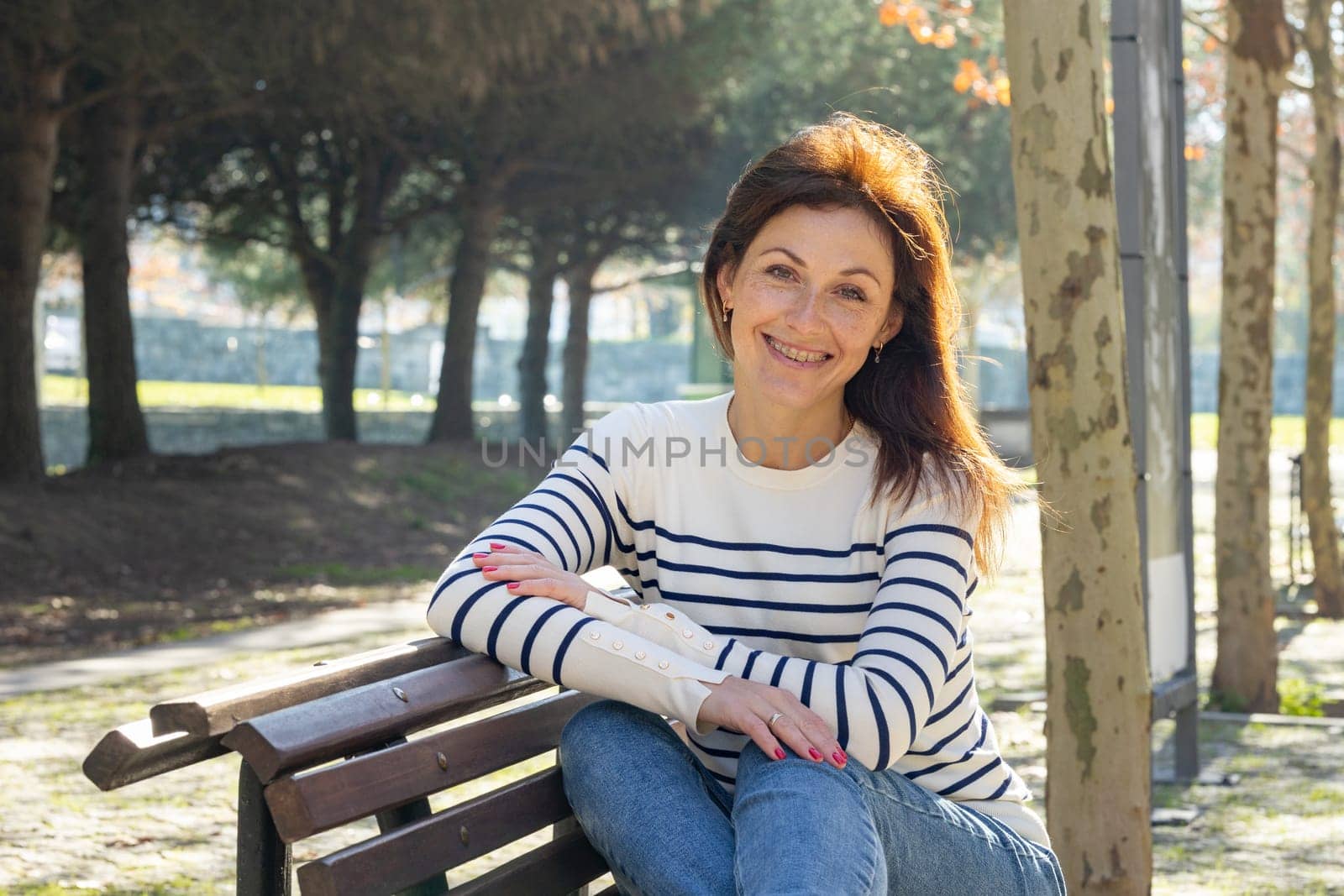A woman wearing dentistry braces is sitting on a bench in a park. She is smiling and wearing a striped shirt