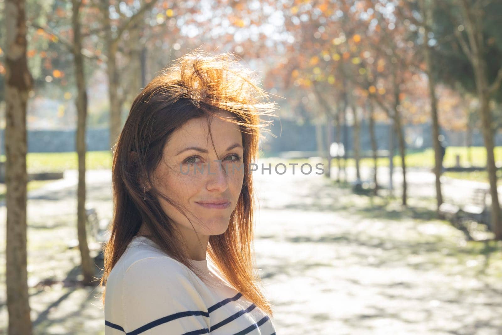 A woman with red hair is standing in a park. She is smiling and looking at the camera