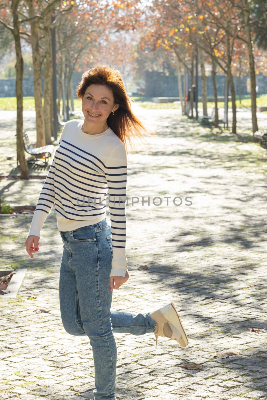 A woman in a striped sweater and blue jeans poses for a picture in a park. She is smiling and she is enjoying herself