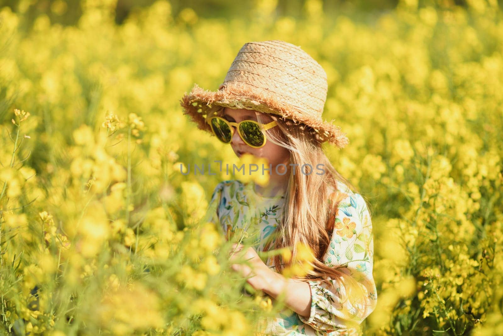 A girl in a dress in a field of rapeseed smelling flowers by Godi