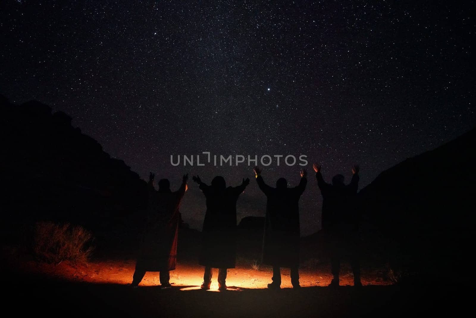 Four persons wearing long warm coats standing in night desert, hands raised to sky with stars, light on the ground, view from behind only silhouettes visible by Ivanko