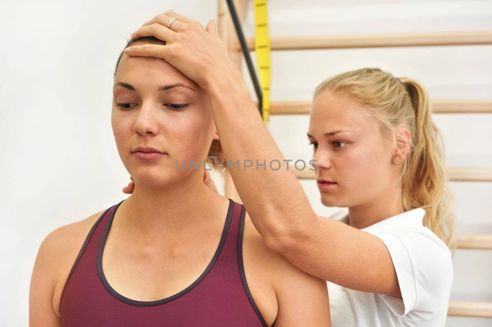 Young physiotherapist exercising with her female patient, fixing head and neck using hand, during exercise