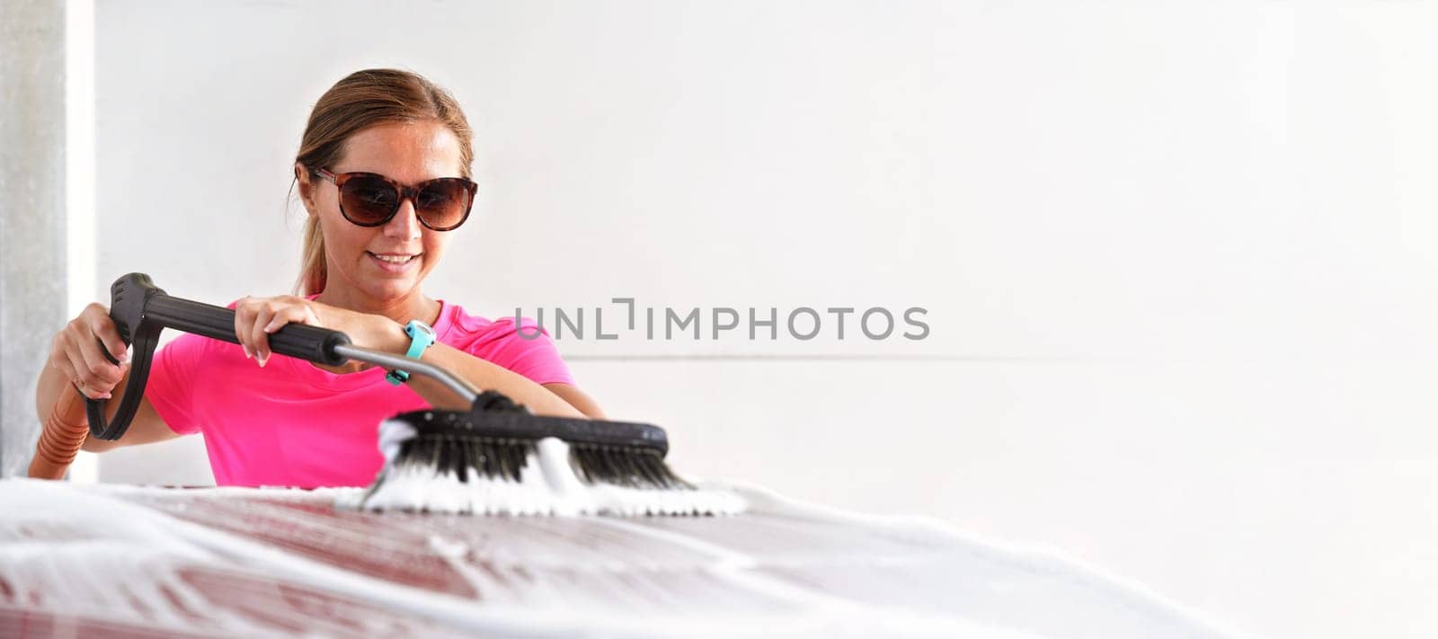 Young woman wearing pink t shirt and sunglasses cleaning her car in self serve carwash, wide banner with space for text right side by Ivanko