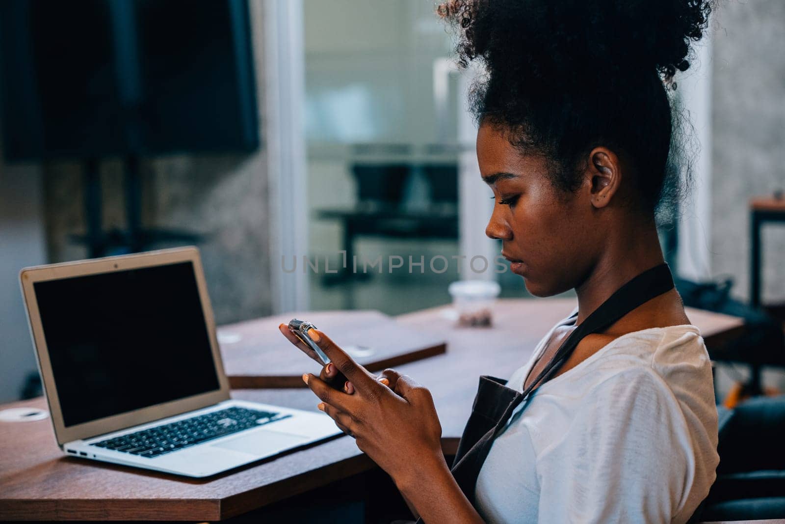 Close up portrait of focused black woman coffee shop owner in apron using smartphone in her cafe. Multi tasking barista manages orders communicates runs her own business.