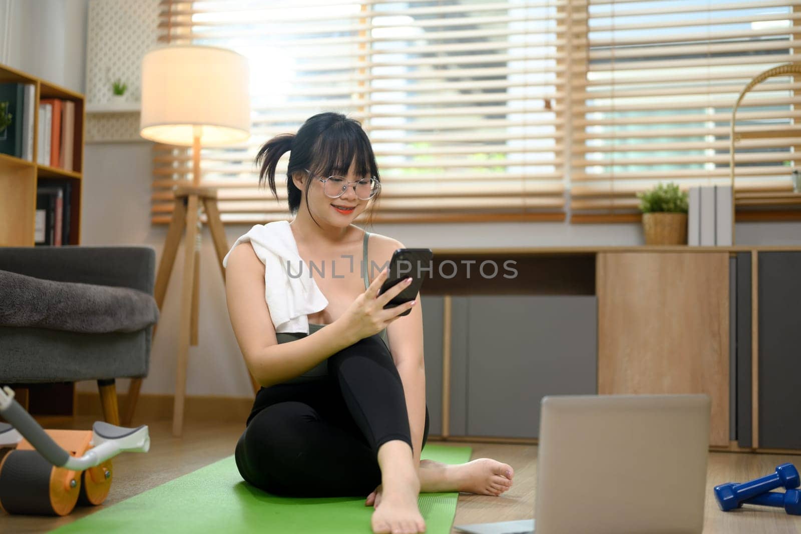 Young woman using smartphone on mat resting after fitness training at home.