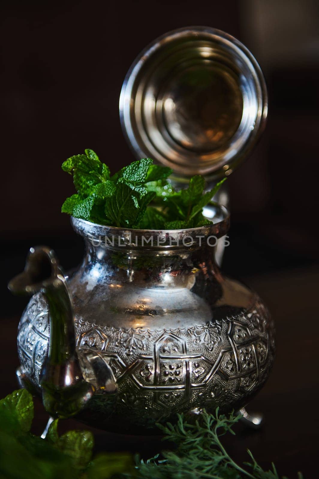 A silver teapot and a bunch of fresh green mint on the foreground. Traditional Moroccan tea as symbol of the hospitality of Moroccan people. Selective focus. Moroccan tea a la menthe