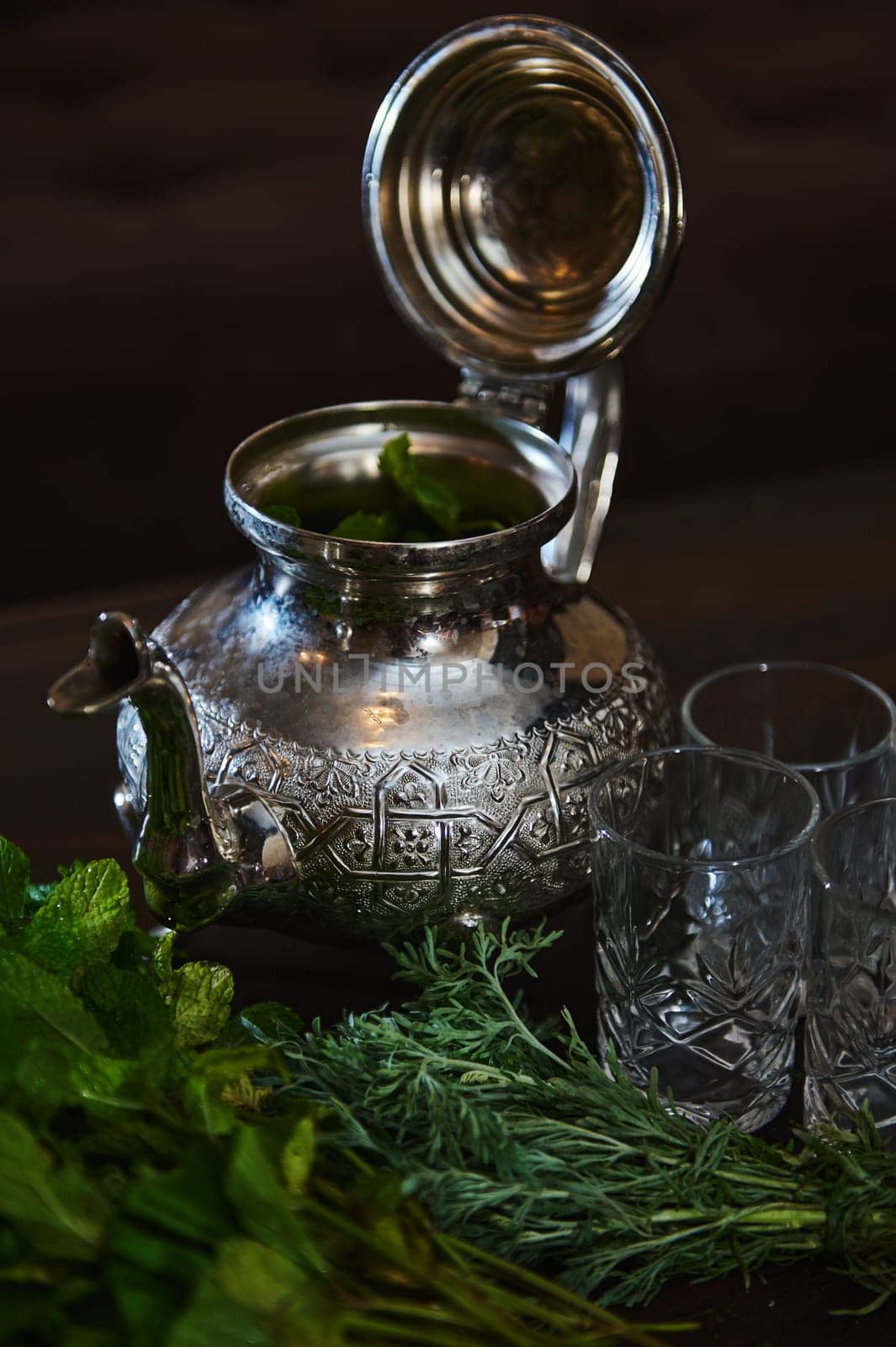 A silver teapot and a bunch of fresh green mint on the foreground. Traditional Moroccan tea as symbol of the hospitality of Moroccan people. Selective focus. Moroccan tea a la menthe