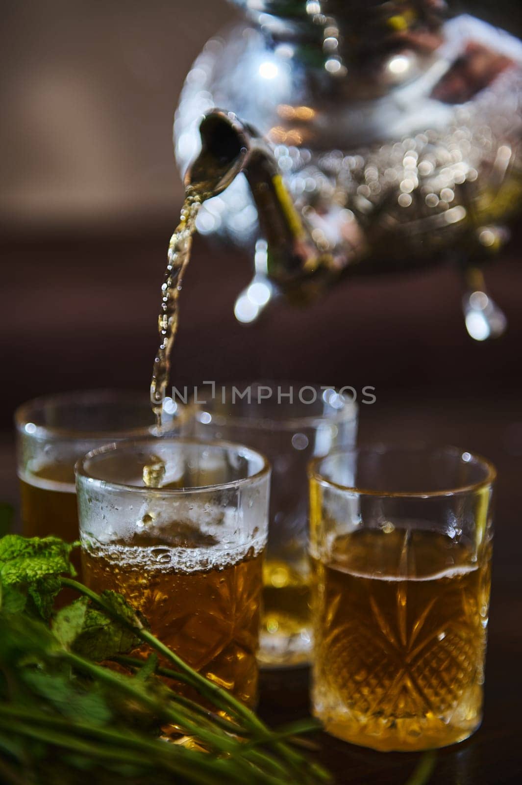 Close-up of pouring delicious Moroccan mint tea from a silver teapot into drinking glasses. Bunch of fresh mint in the foreground. Tea break in Morocco