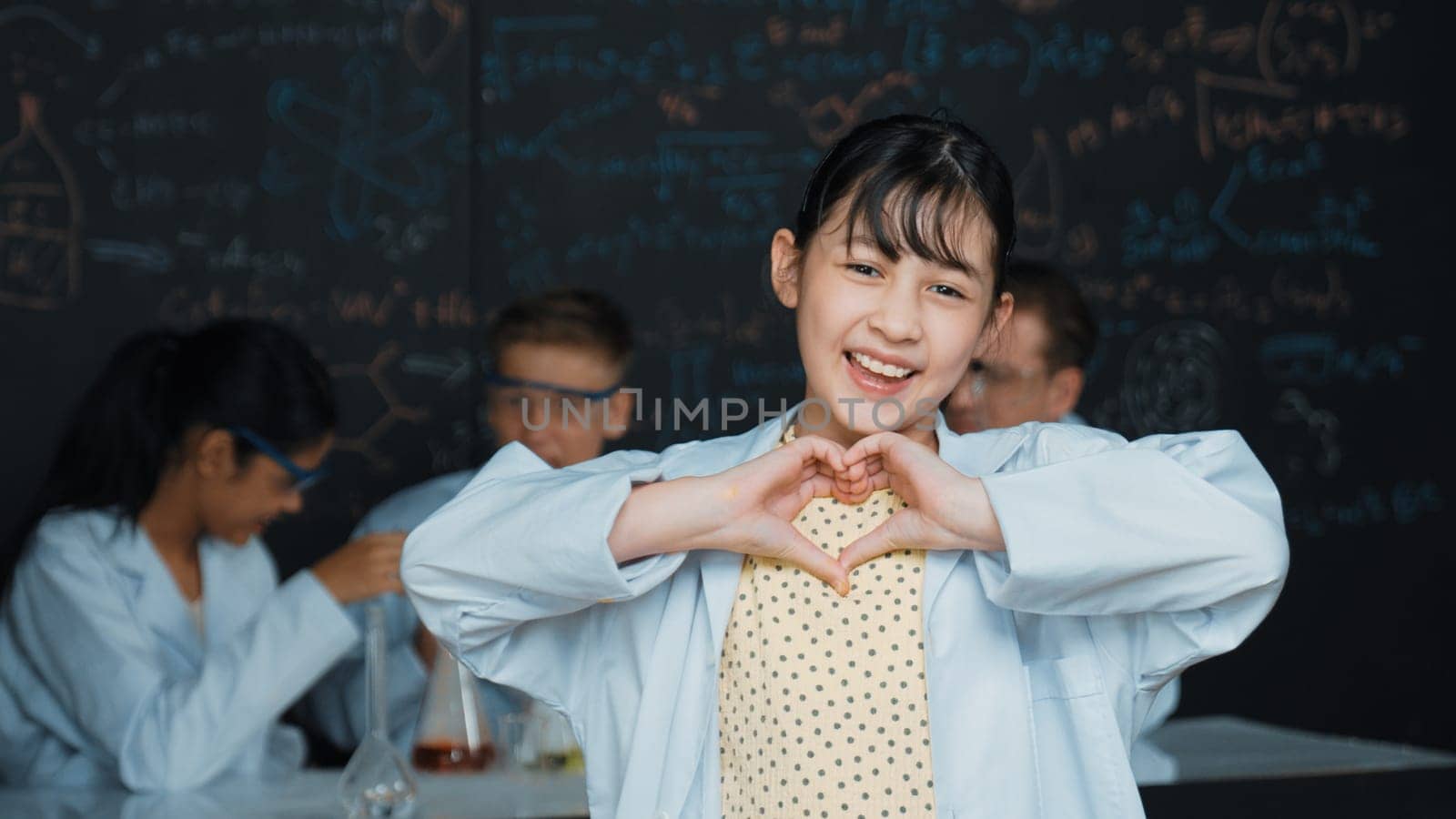 Asian scientist show miniheart to camera while diverse group doing experiment. Young schoolgirl standing at blackboard with chemical theory written while wearing lab coat at laboratory. Edification.