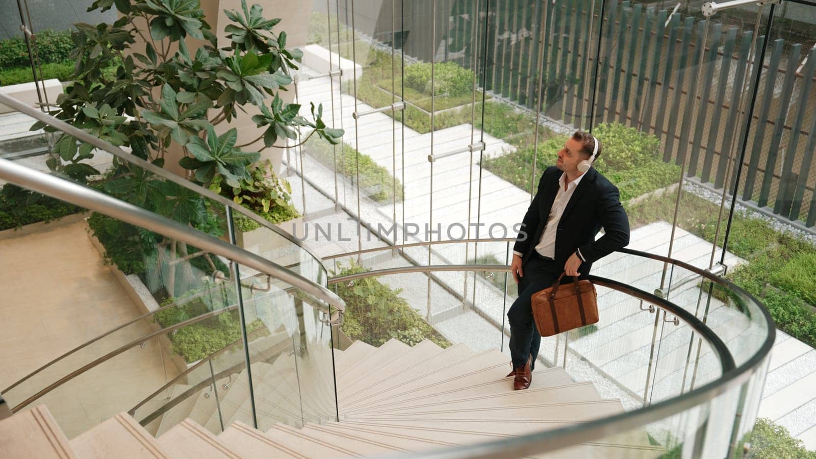 Top view of caucasian businessman listening relaxing music wearing headphone while standing at spiral stair cases at modern building. Project manager enjoy listening song while move to rhyme. Urbane.