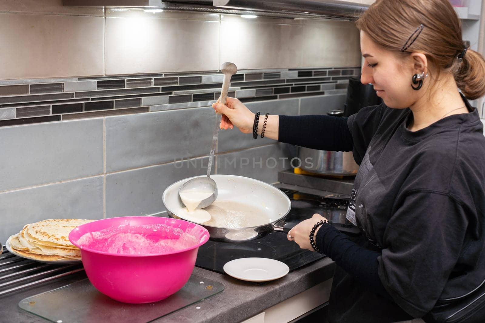 In the kitchen, a girl is standing by an induction cooker, pouring the ready-made pancake batter into a hot pan. Homemade pancakes from a woman's hand.