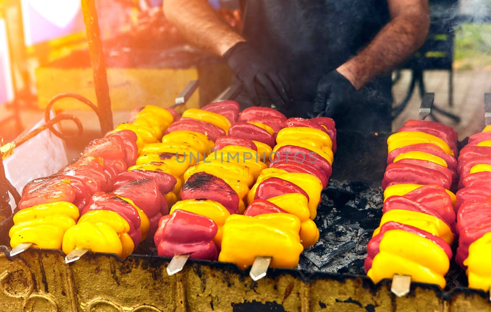 Rows of red, yellow, and orange bell peppers on skewers being grilled outdoors at a food market. by Hil