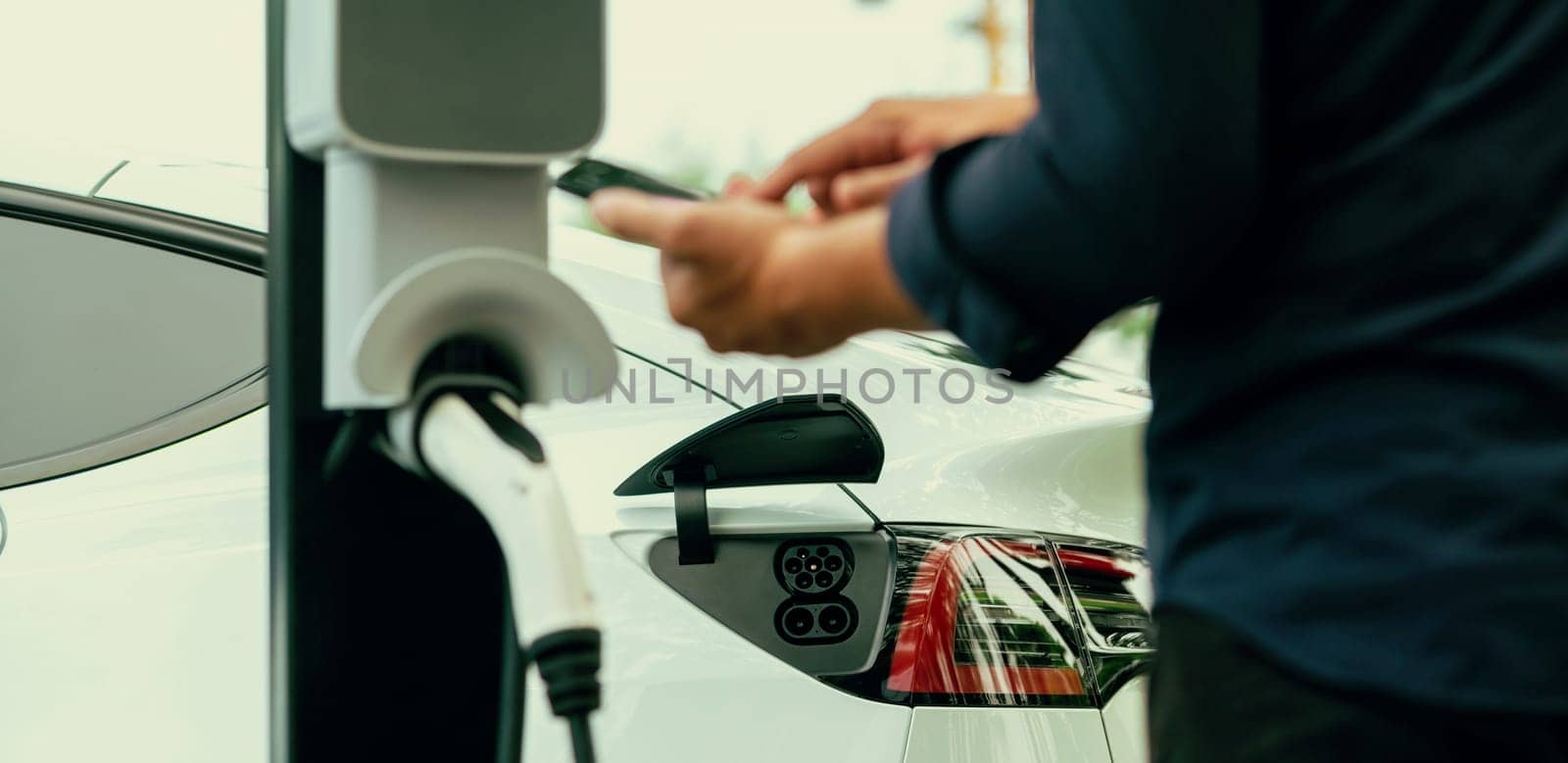 Man using smartphone online banking application to pay for electric car battery charging from EV charging station during vacation road trip at national park or summer forest. Panorama Exalt