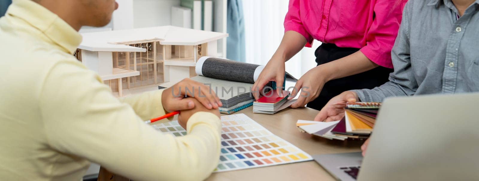 Skilled interior design team carefully selecting curtain materials while coworker selecting the color of curtain with house model placed on meeting table. Creative design concept. Variegated.