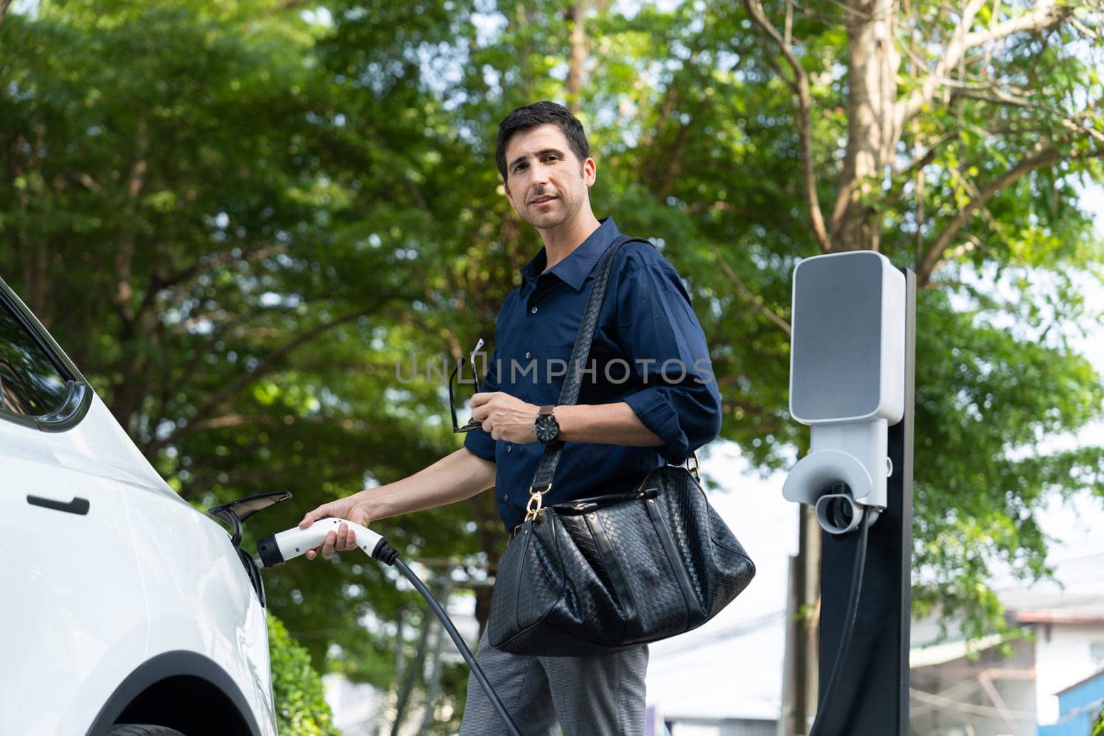 Young man recharge electric car's battery from charging station in outdoor green city park in springtime. Rechargeable EV car for sustainable environmental friendly urban travel lifestyle. Expedient