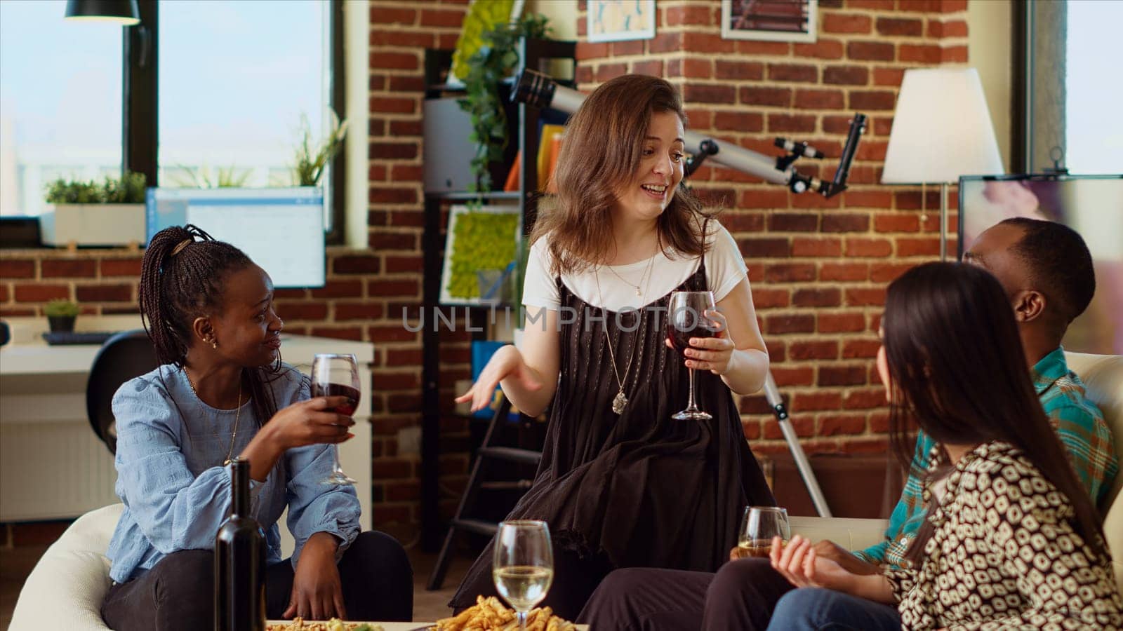 Caucasian woman entertaining friends at house party, sharing with them interesting story. Guest making inclusive group of people laugh in living room while enjoying drinks and charcuterie platter