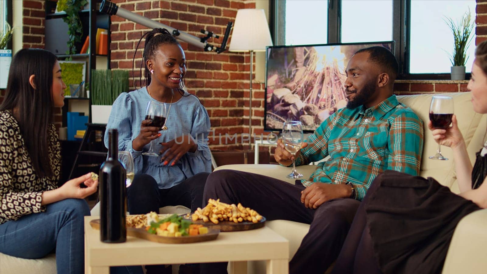 Diverse mates having engaging conversation during apartment party in cozy house. Happy group of people talking in living room, eating snacks and drinking alcoholic beverages