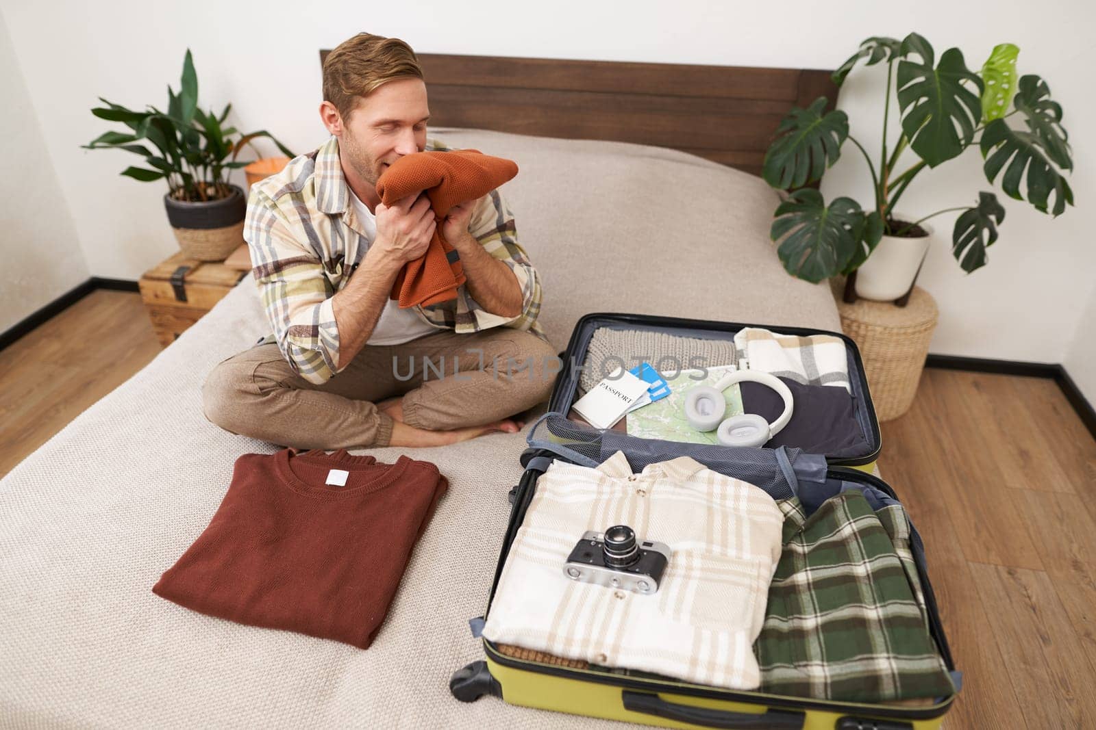 Portrait of happy young man, smells his clothes with pleased face, sits on bed with suitcase, goes on holiday, tries new detergent. People concept