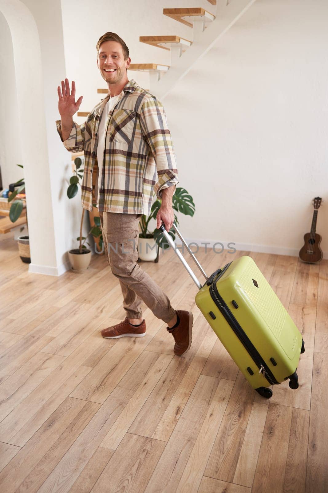 Vertical portrait of happy guy, tourist leaving the apartment with suitcase, waving hand at camera, saying hello or goodbye.