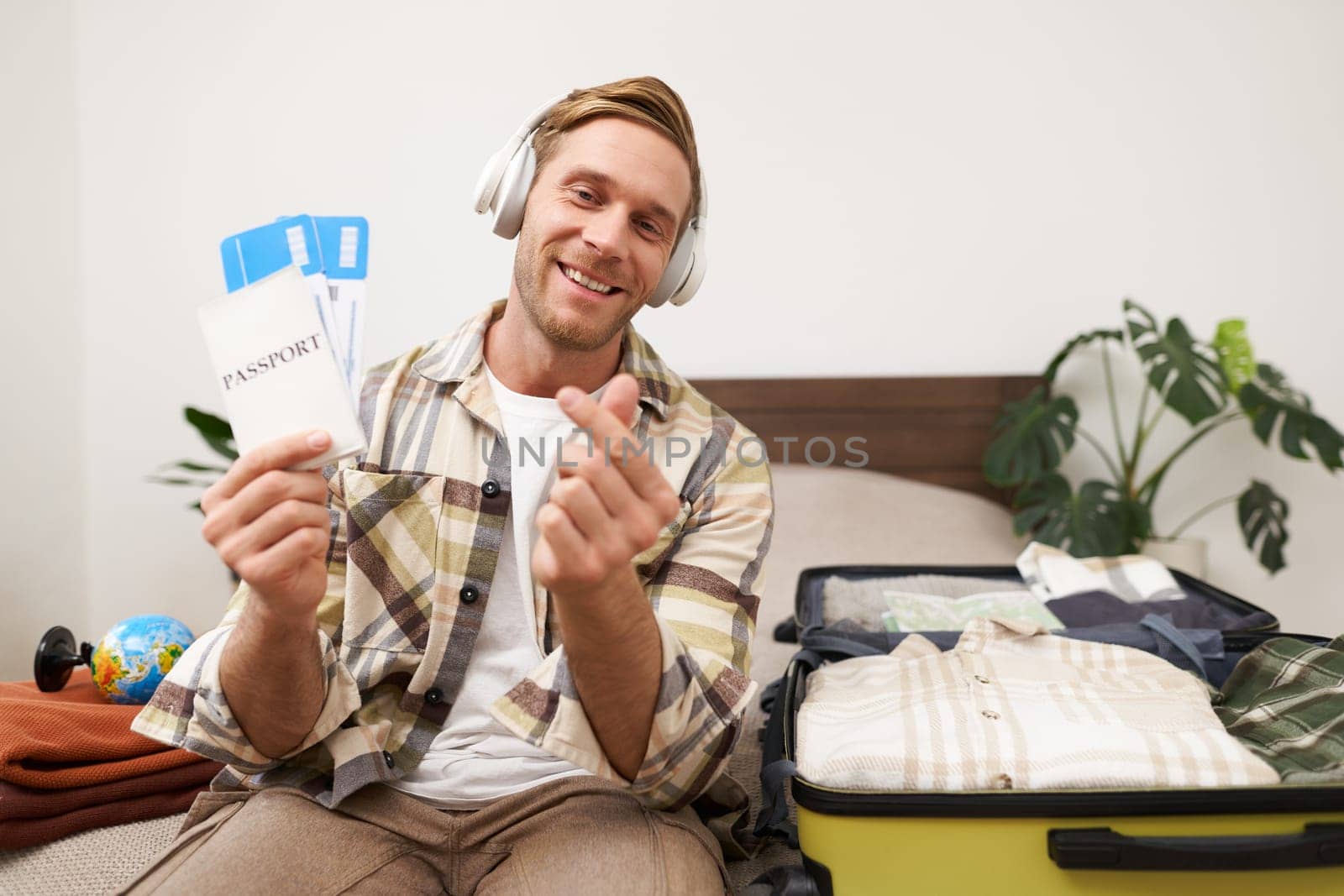 Image of handsome young man, tourist shows two plane tickets and passport, travels on holiday, goes on vacation, packs suitcases by Benzoix