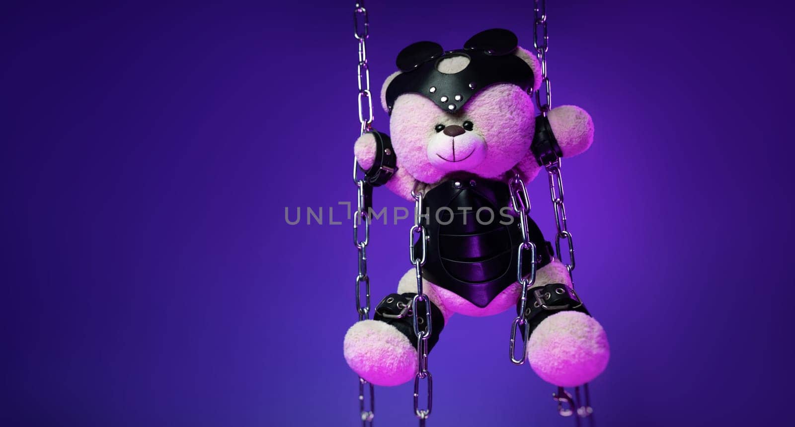 teddy bear in a mask is suspended from chains in leather handcuffs and accessories for bdsm games and sex in neon light against a background of copy paste