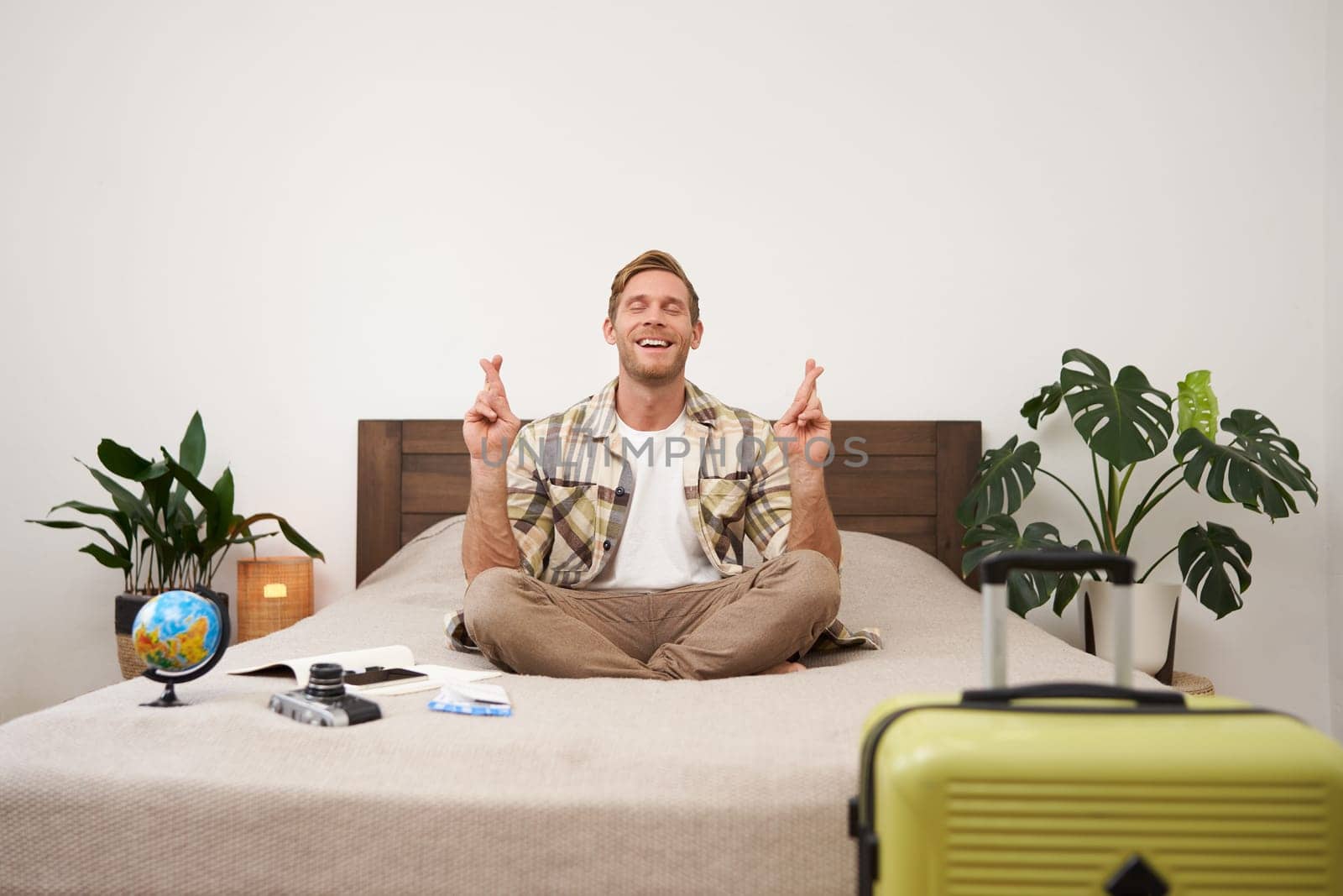 Young man with globe and camera, sits on bed and meditating, has packed suitcase standing near him, going on vacation, praying for good weather on holiday, making wish with fingers crossed.