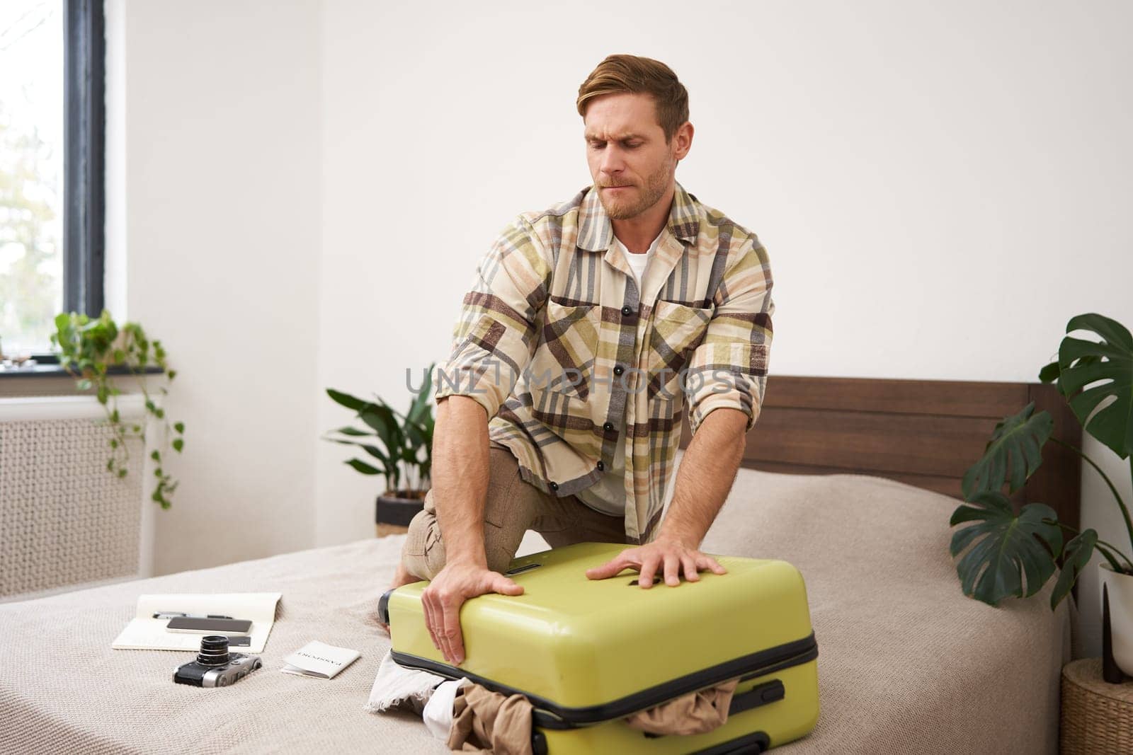 Portrait of young man, tourist packs his bag, closing full suitcase, trying to push the luggage inside to zip the bag, sitting on bed in bedroom. Tourism and adventure concept