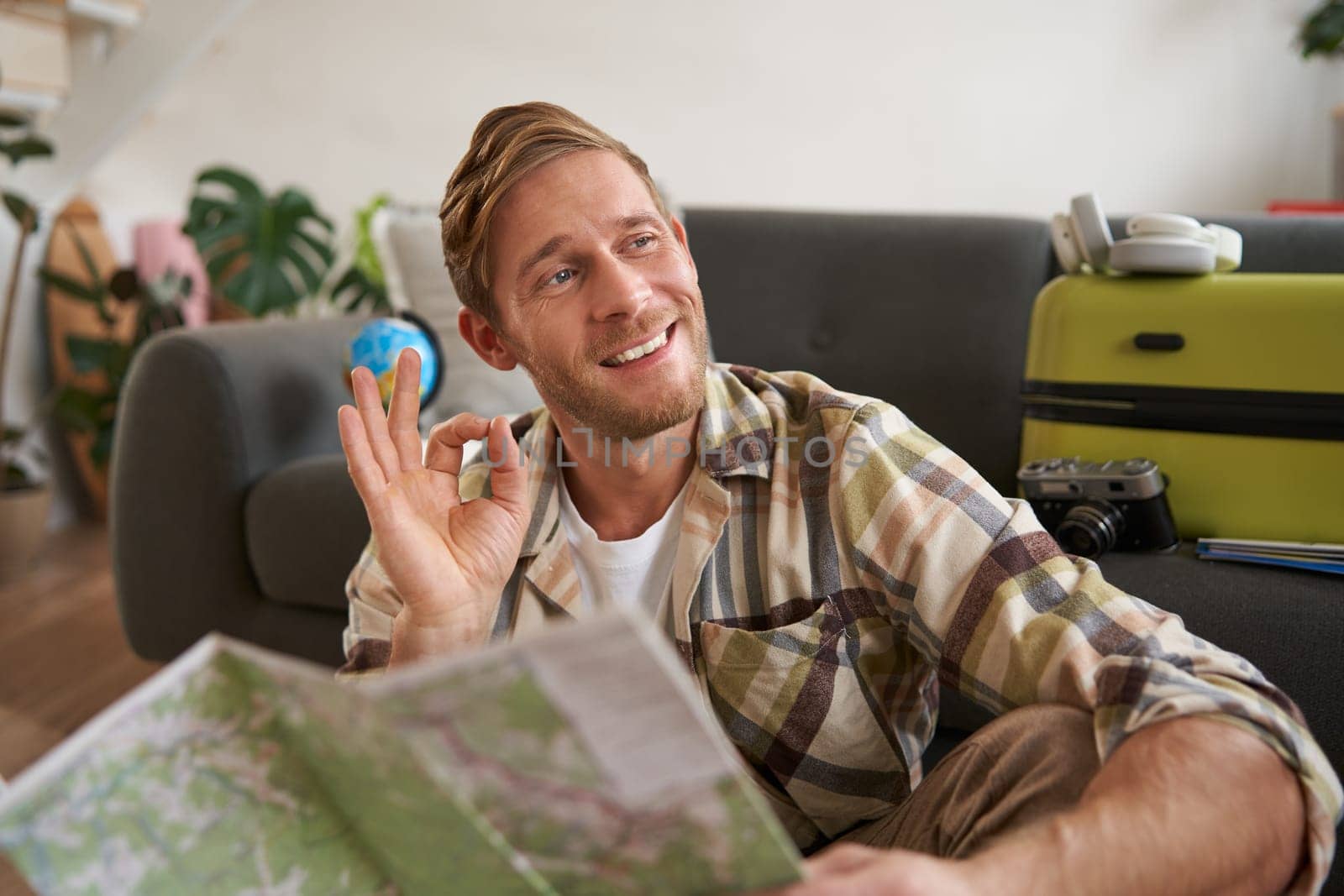 Portrait of smiling handsome guy with travel map, plans tour, shows okay, ok hand gesture, sits on floor with packed suitcase. Journey, travelling and tourism concept