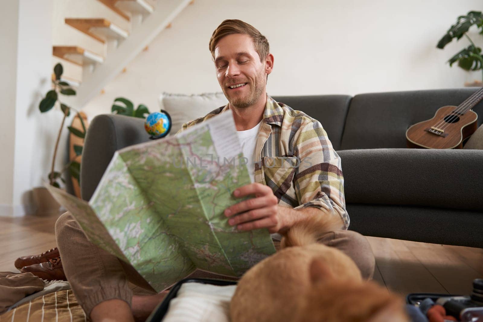 Portrait of young man, traveller, sitting packing suitcase, looking at travel map, choosing holiday destination, studying the route on vacation. Tourism concept