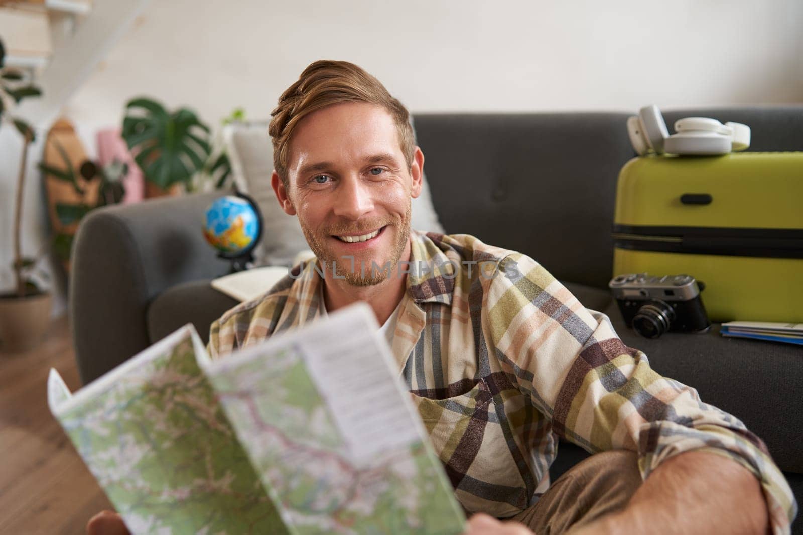 Portrait of handsome smiling man, holding map, sitting with suitcase in living room, going abroad on holiday, studying his route. Tourism concept