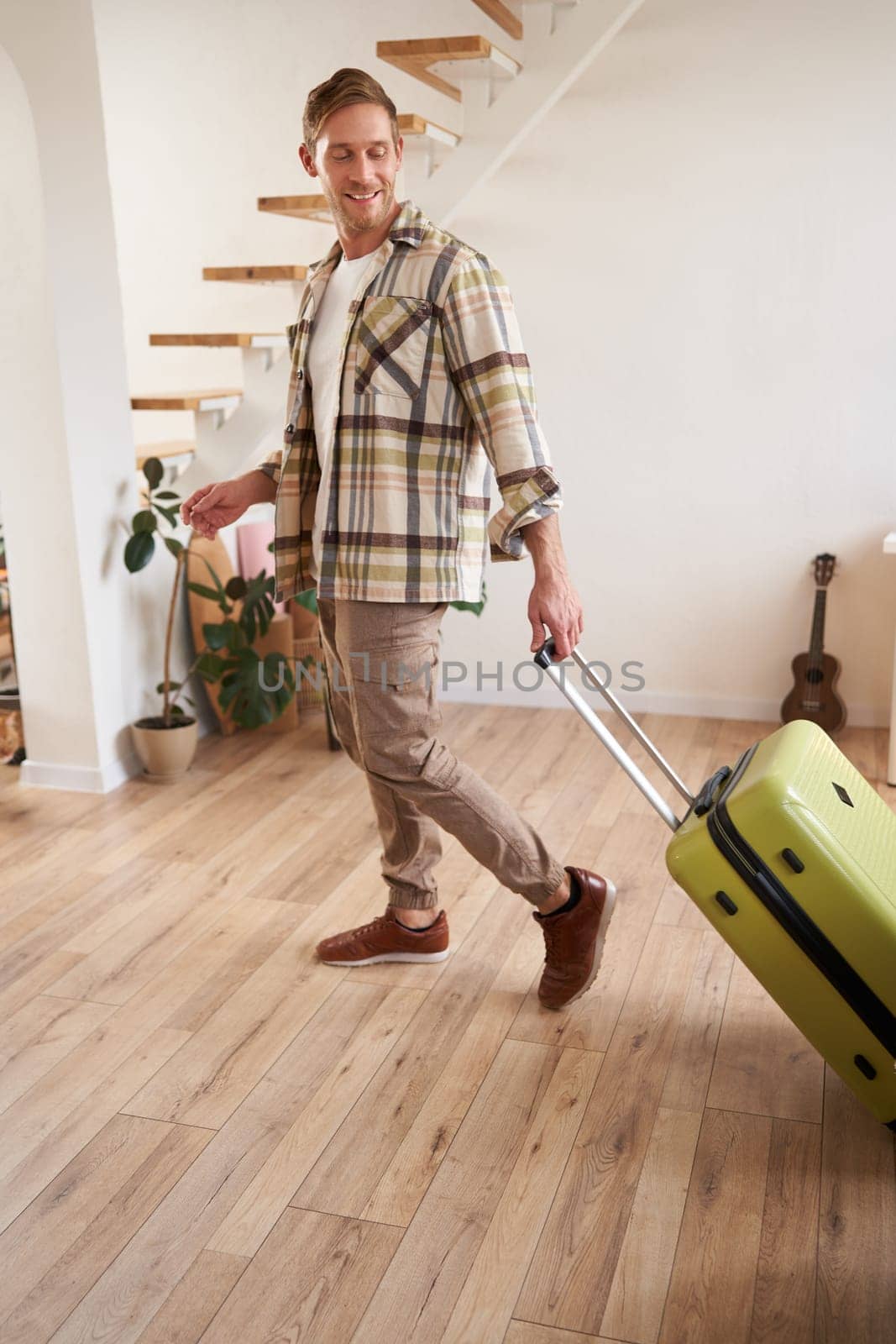 Vertical portrait of happy young man with suitcase, walking indoors, leaving a flat with luggage, going on business trip or vacation. Copy space