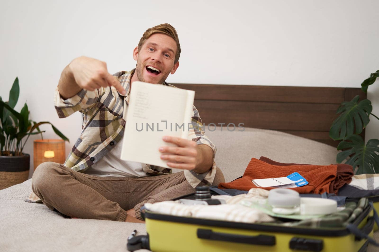 Portrait of young man pointing at his list of items, packing clothes on vacation, showing notebook, sitting near opened suitcase. Lifestyle and people concept