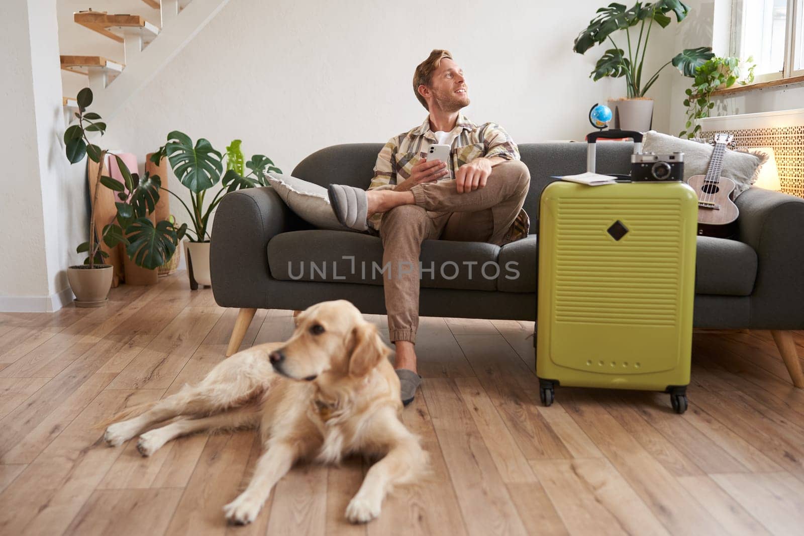 Portrait of happy, handsome young man with suitcase, sitting with his dog in living room, prepared to go on vacation, going on holiday. Tourism concept
