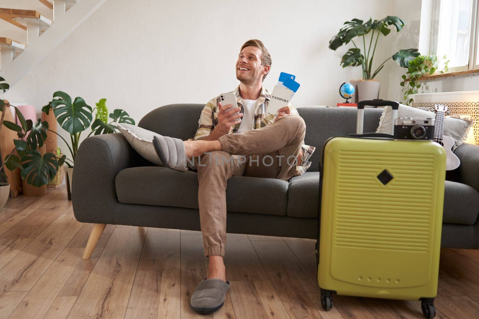 Handsome young man sits on sofa with suitcase, holds smartphone, books hostel or holiday vacation, chats on mobile phone. Tourism and travelling concept
