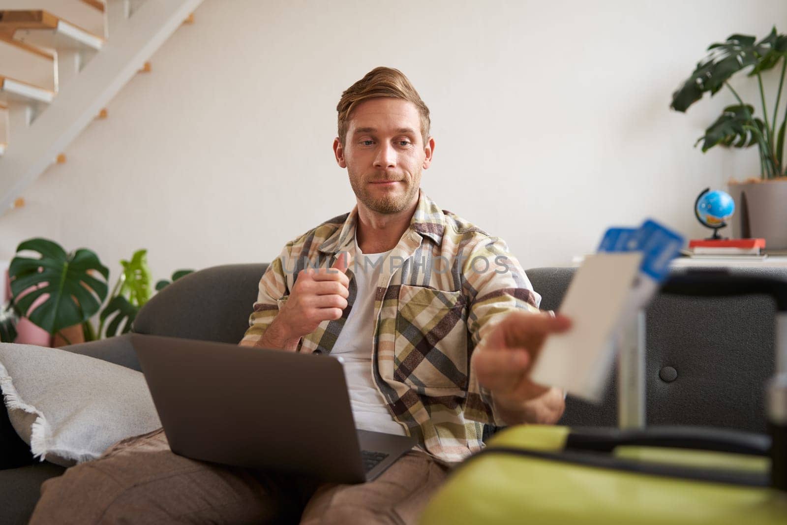 Portrait of happy young man booking holiday for two, has plane tickets, looks at laptop, sits in living room with computer by Benzoix