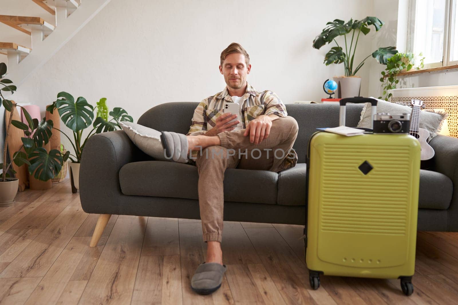 Portrait of man sits with mobile phone in living room, packed suitcase, chats on smartphone app, looks for holiday destinations, goes on vacation.